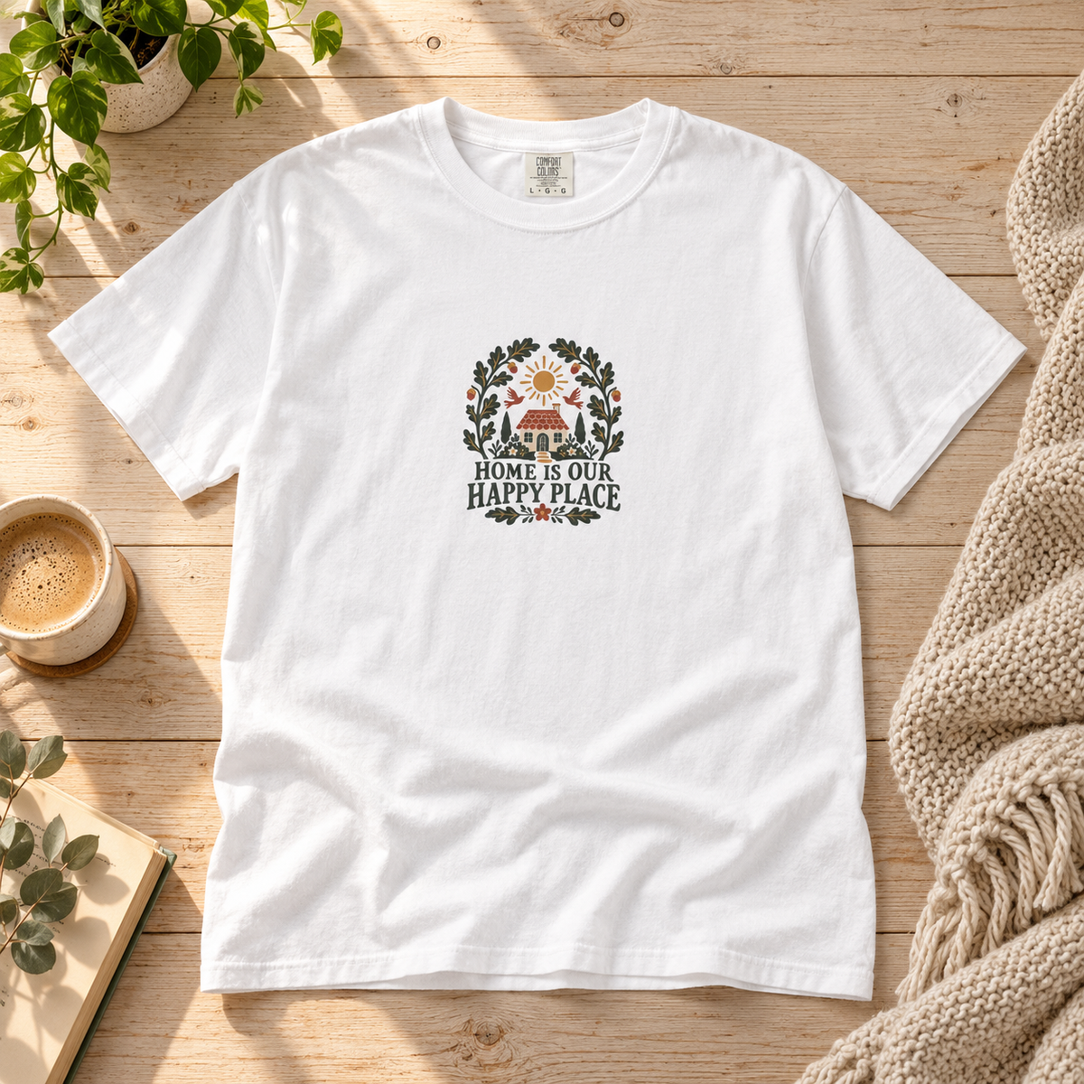 Lifestyle mockup of the white Home Is Our Happy Place graphic T-shirt on a pale wood table with coffee and a neutral knit throw.