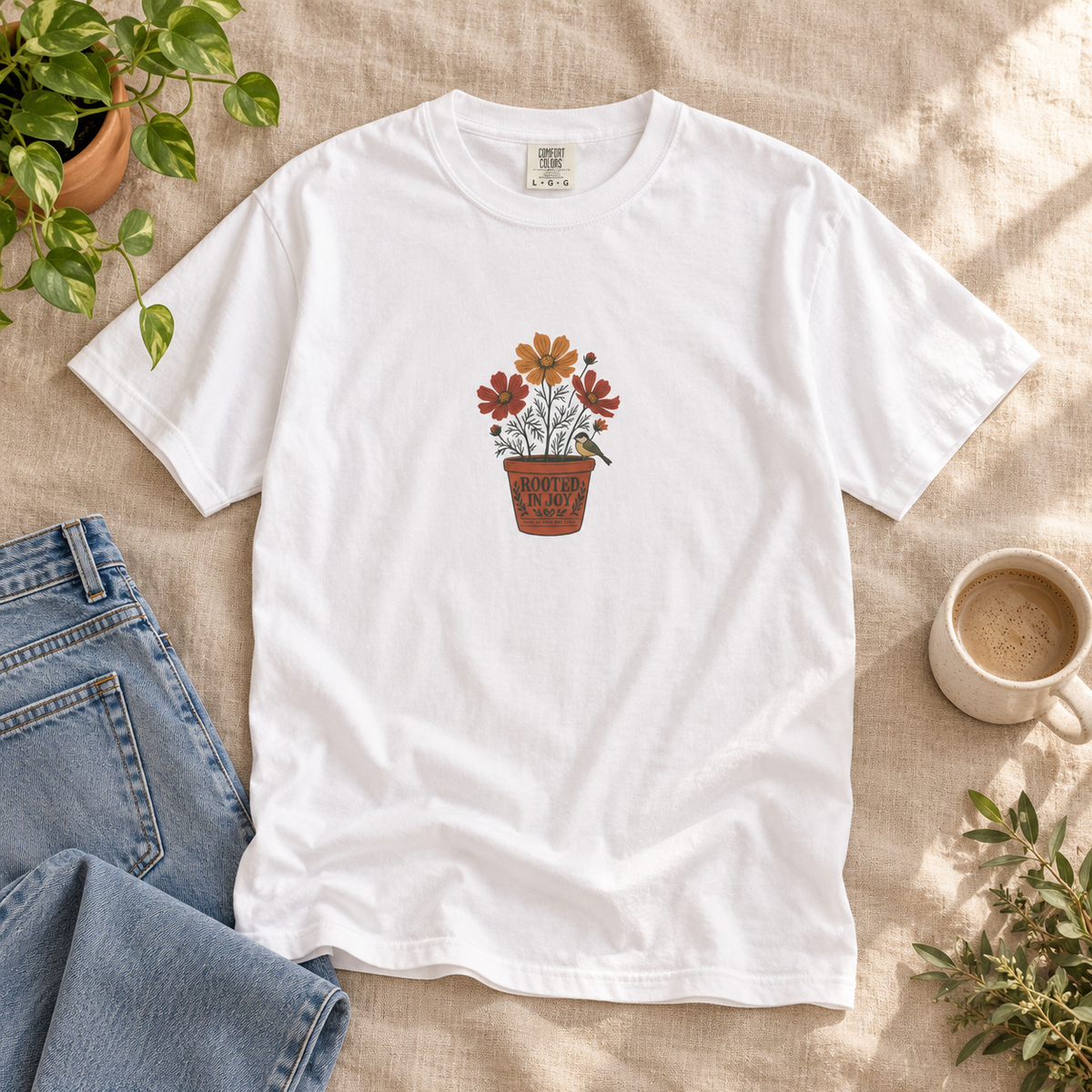 Lifestyle mockup of the white Rooted in Joy botanical T-shirt on beige linen with jeans, a plant and coffee.